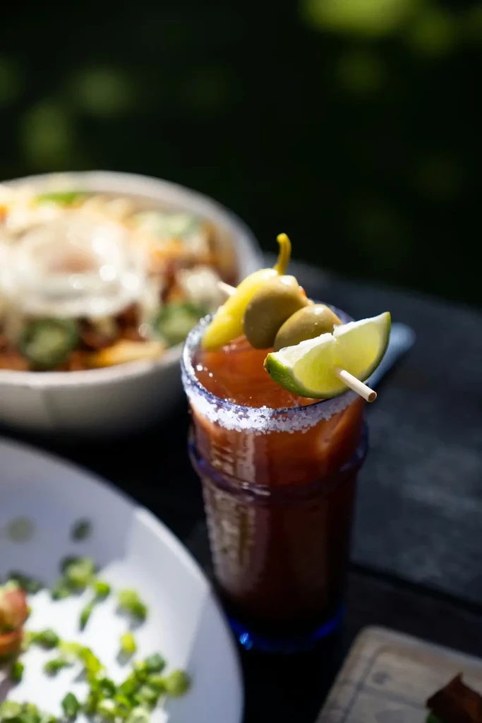 Bloody Mary cocktail in dark glass, garnished with lime, olives, pepper.