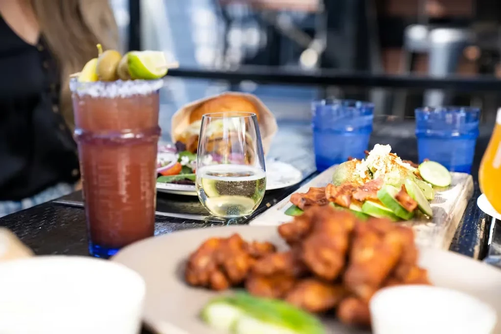 Restaurant table with chicken wings, a Bloody Mary, wine, and a burger.