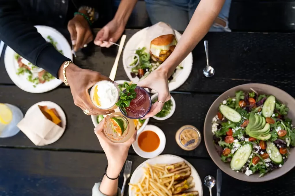 Hands toast drinks over plates of salad, burgers, and fries on a table.