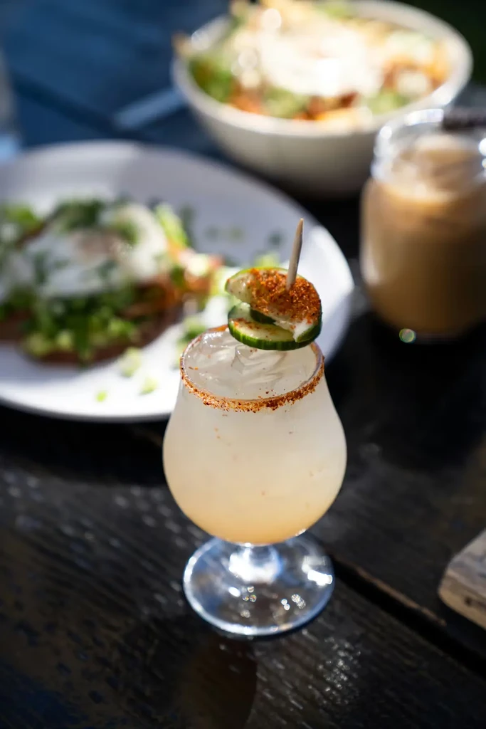 Frothy drink in stemmed glass, sugared rim, garnished with cucumber and lime.