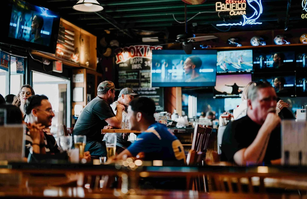 Lively sports bar interior with blurred patrons, multiple TVs, and Pacifico sign.