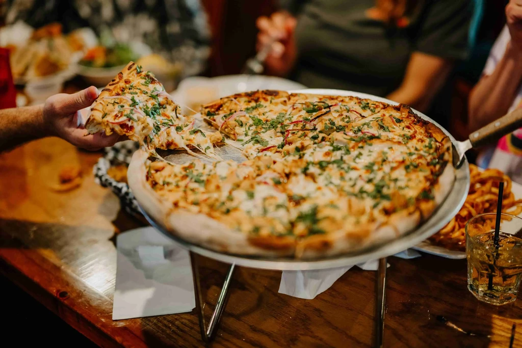 Hand pulling a cheese-stringed slice of chicken pizza from a stand.