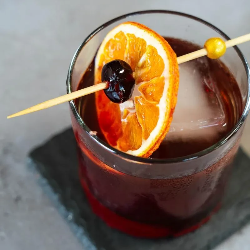 Reddish-brown cocktail, old-fashioned glass, ice, dried orange, and cherry.