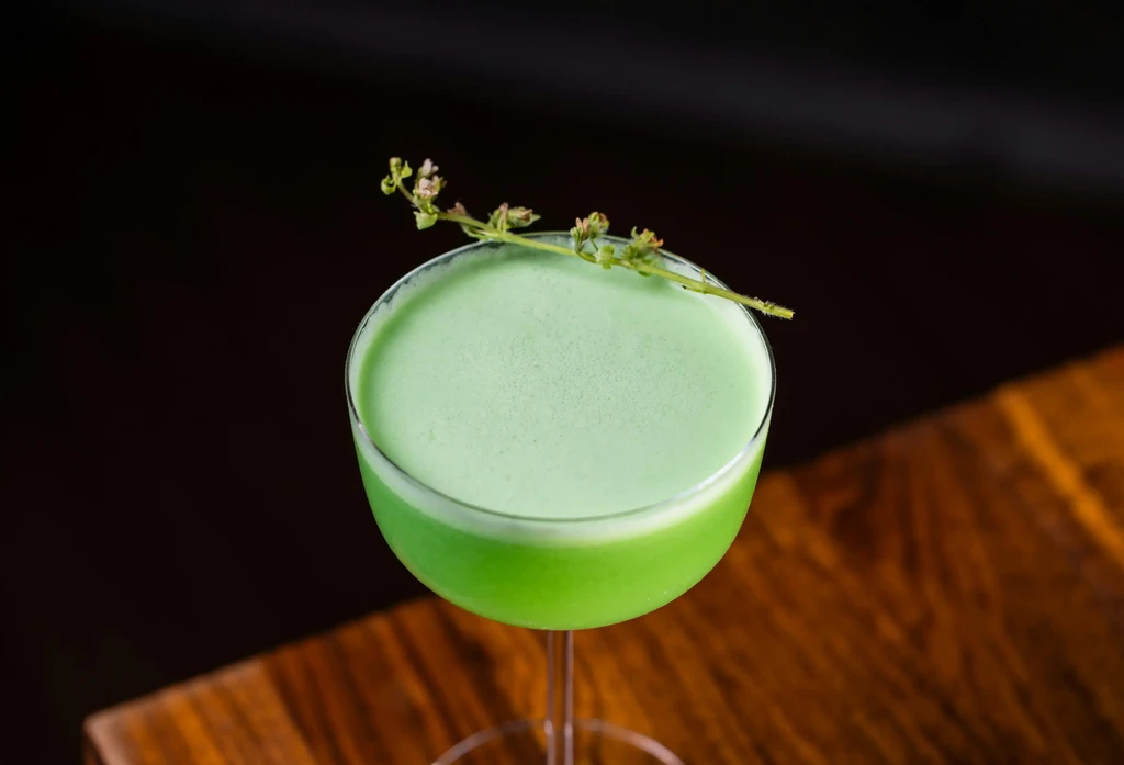 Green cocktail in coupe glass, frothy top, garnished with a sprig.