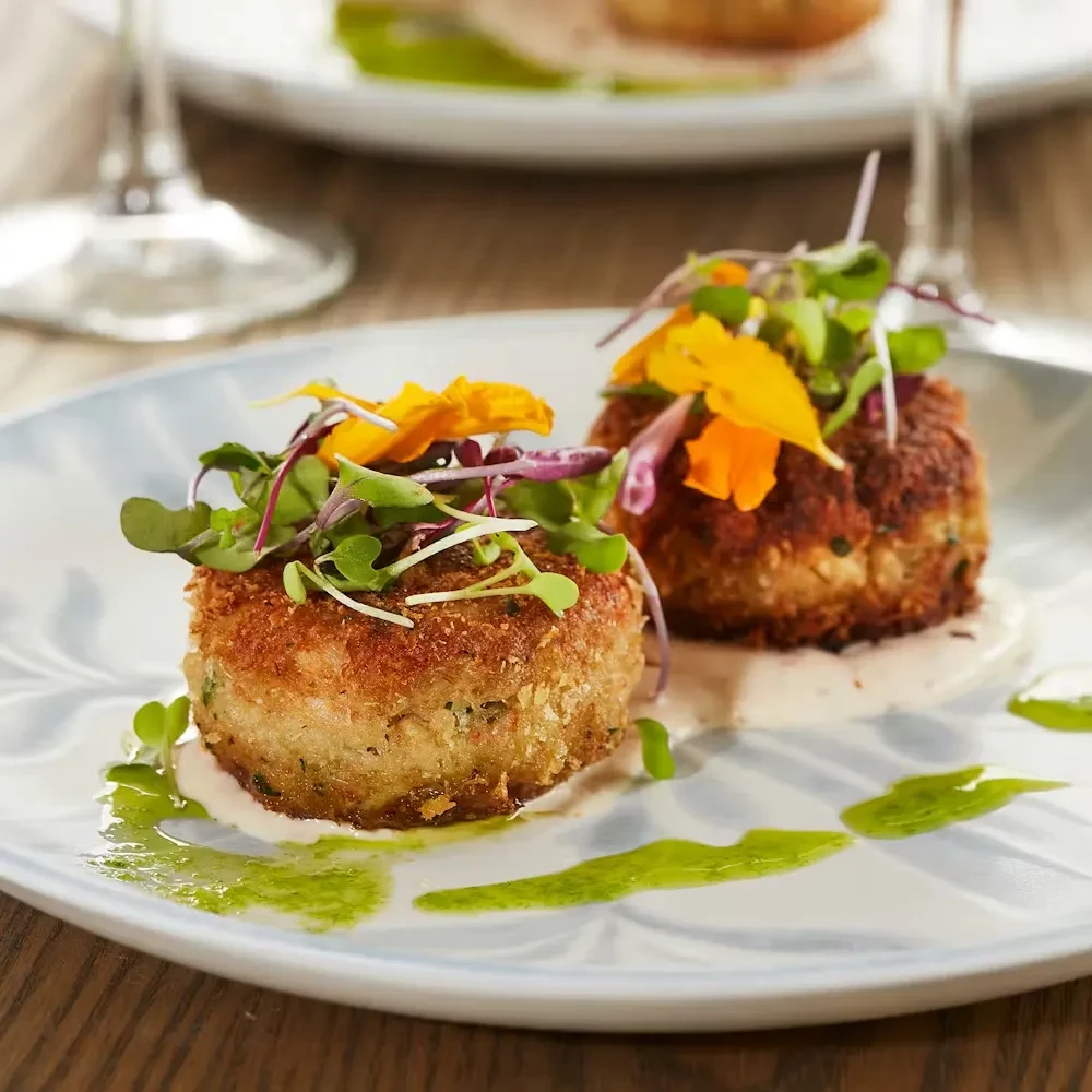 Two crispy crab cakes on a blue plate, garnished with microgreens and flowers.