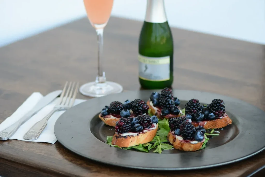Five berry crostini, cutlery, and pink sparkling wine on a table.