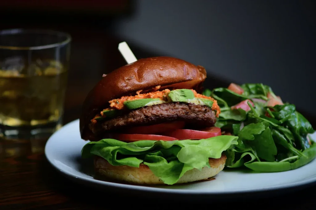 Gourmet hamburger with avocado, side salad, and drink on white plate.