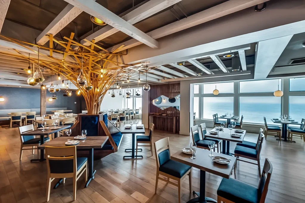 Modern restaurant with lit wooden tree sculpture, ocean view, and fish art.