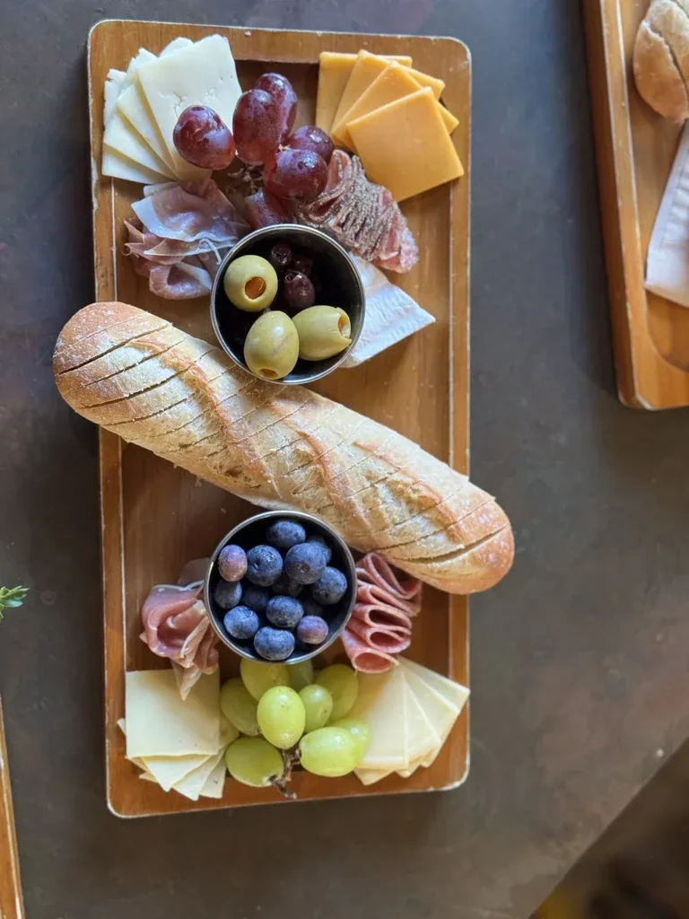 Arranged wooden charcuterie board with baguette, cheese, meats, fruits, and olives.