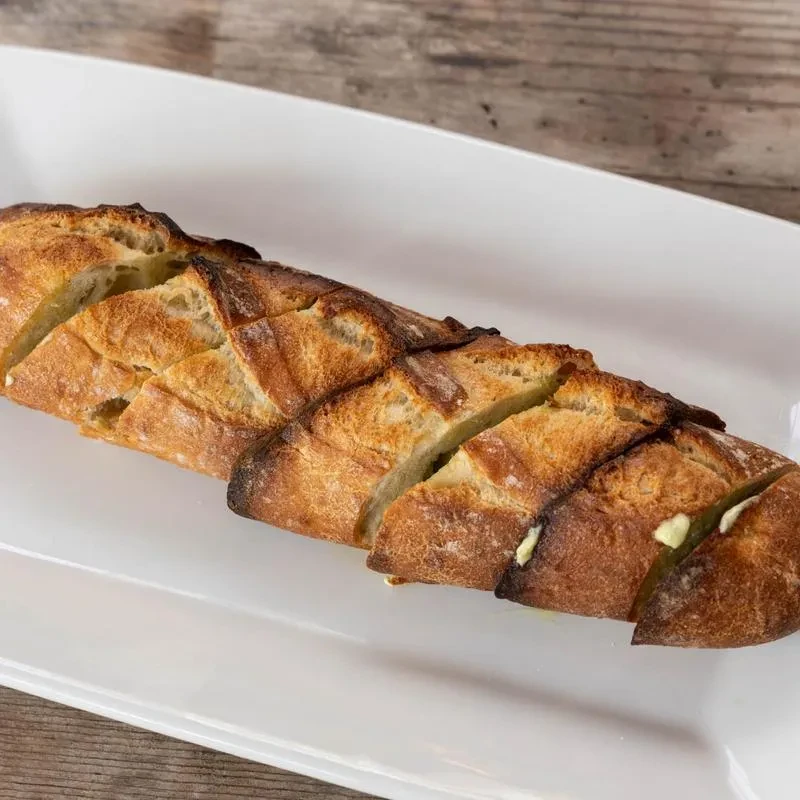 Partially sliced, golden baguette with melted butter on a white dish.