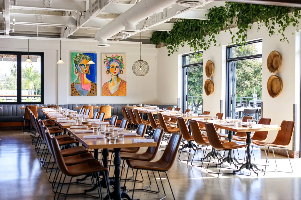 Bright, empty modern restaurant with set tables, abstract art, and foliage.