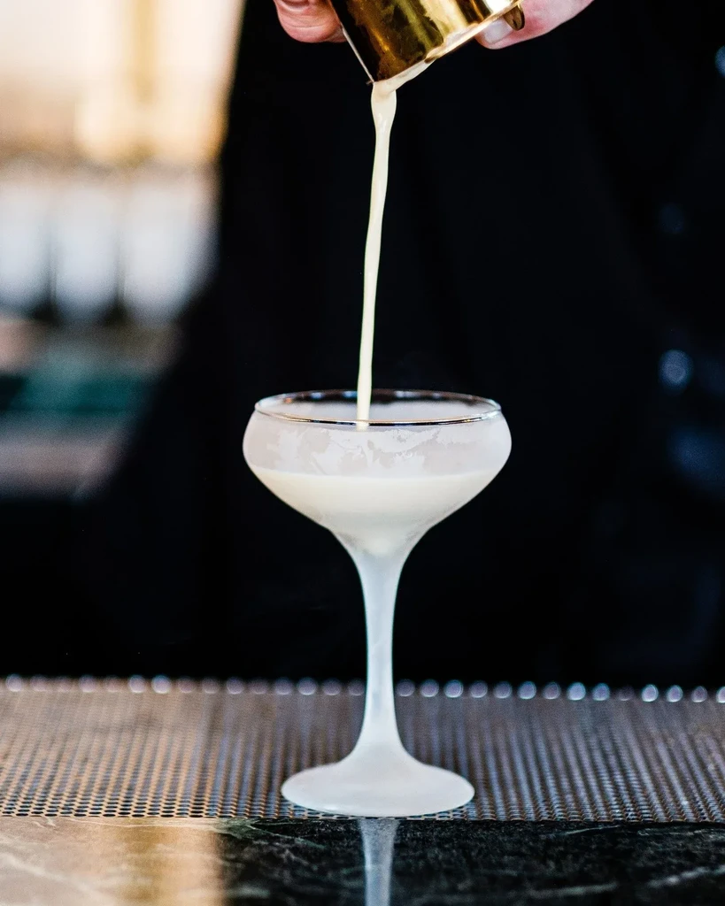 Pouring creamy white cocktail from golden shaker into frosted coupe.