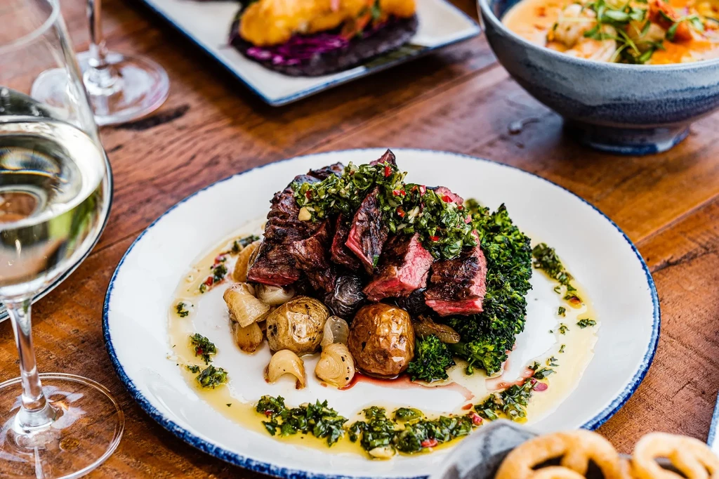 Sliced medium-rare steak with chimichurri, roasted potatoes, and broccoli on a rustic table.