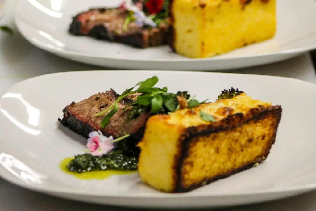 Two artfully plated dishes with meat, gratin, greens, and flower.