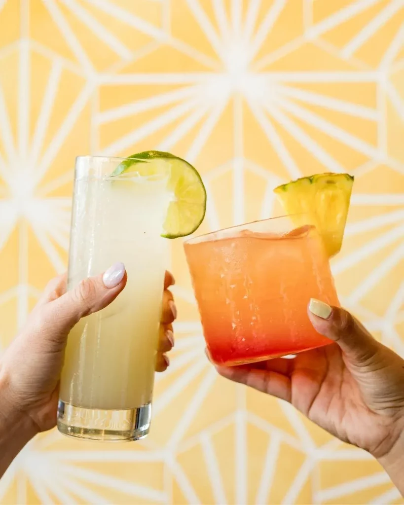 Two hands hold different cocktails with lime and pineapple on yellow.