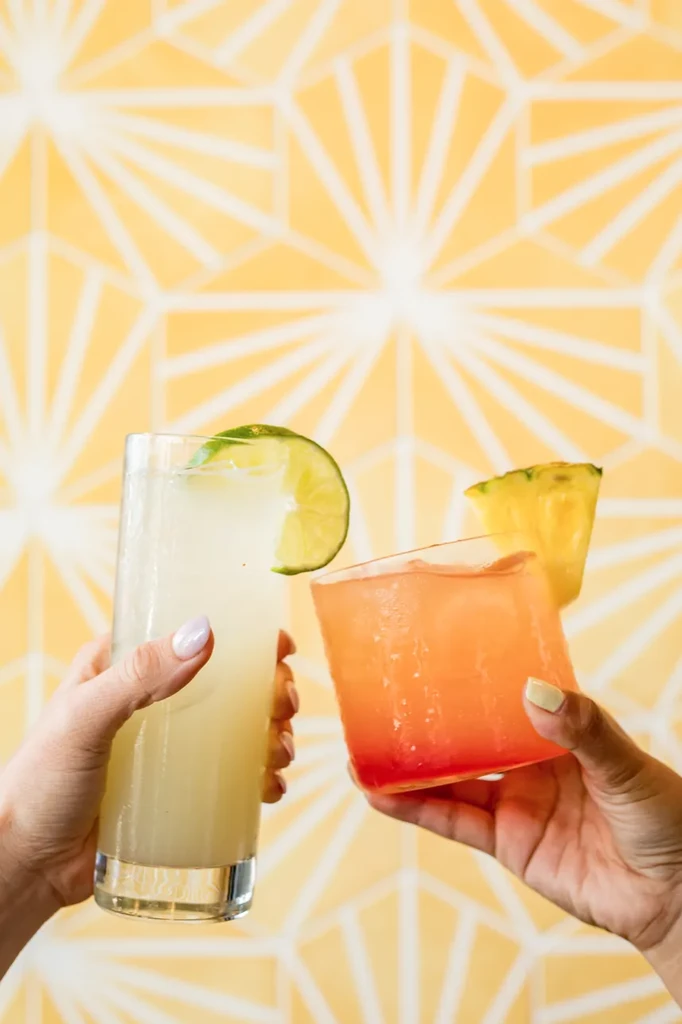 Two hands holding different iced cocktails with lime and pineapple.