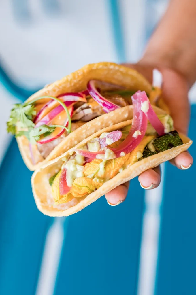 A hand holds two colorful tacos, one with squash blossoms.