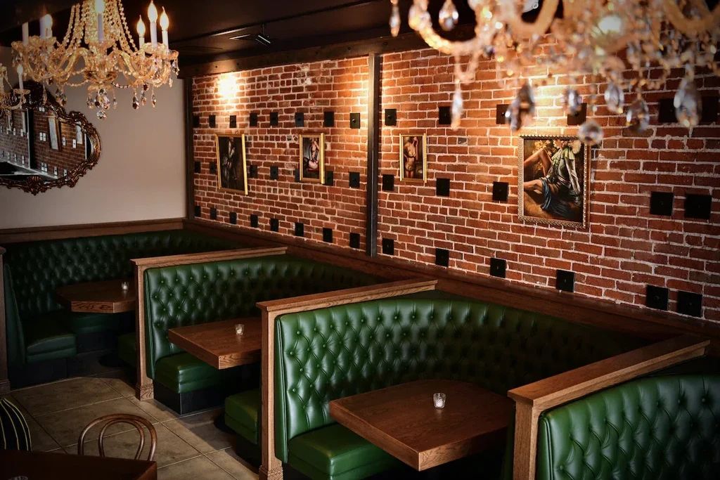 Restaurant interior with tufted green booths, brick wall, and chandeliers.