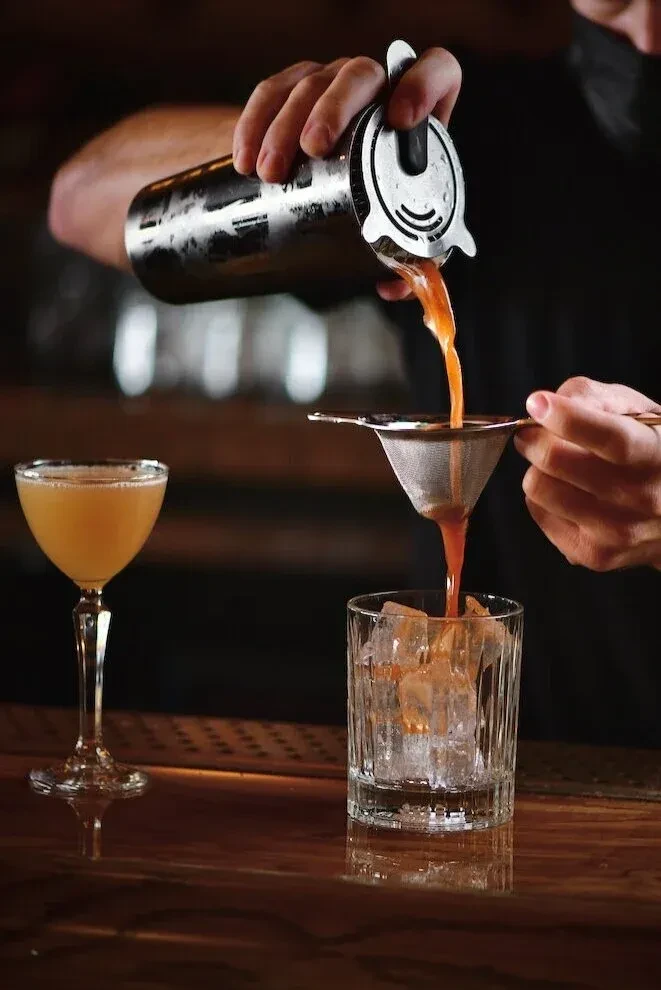 Bartender's hands strain orange cocktail from shaker into iced glass.
