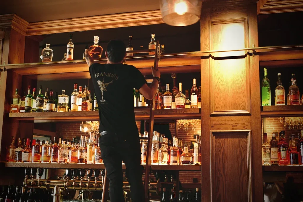 Bartender on ladder organizes bottles on dark wood liquor shelves.