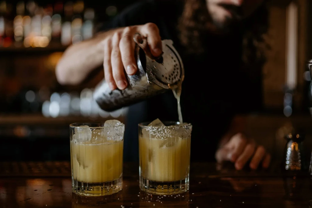 Close-up: Bartender pours yellow cocktail from shaker into salt-rimmed glass.