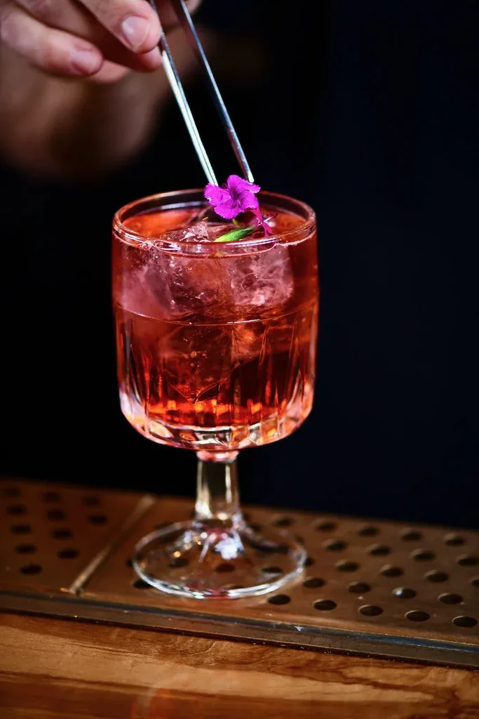 Reddish-orange cocktail in a cut-glass goblet, garnished with a fuchsia flower.