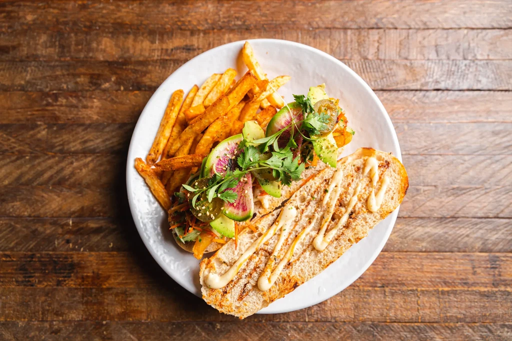 Overhead: Sandwich with avocado, radish, cilantro, fries on a wooden table.