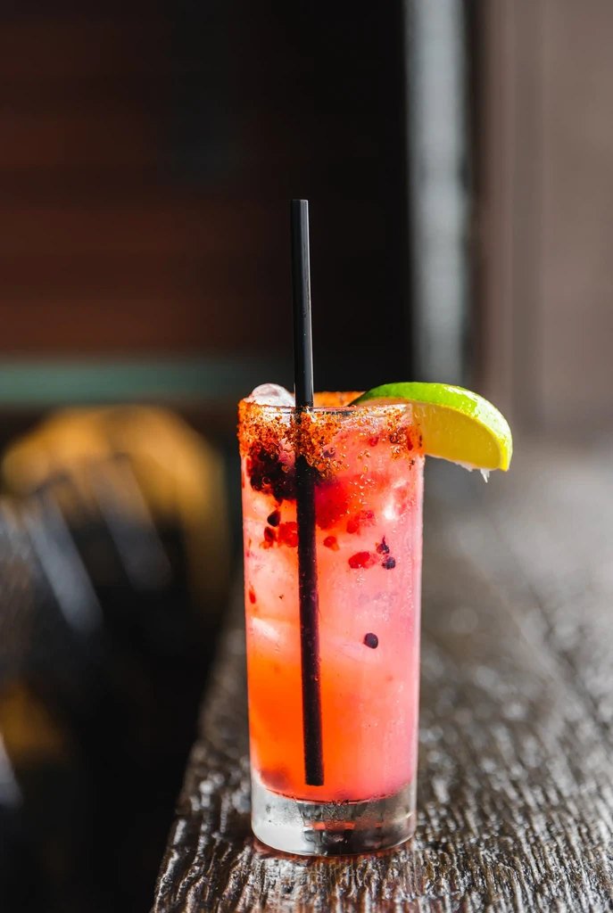 Reddish-pink cocktail with ice, seeds, lime wedge, and seasoned rim.
