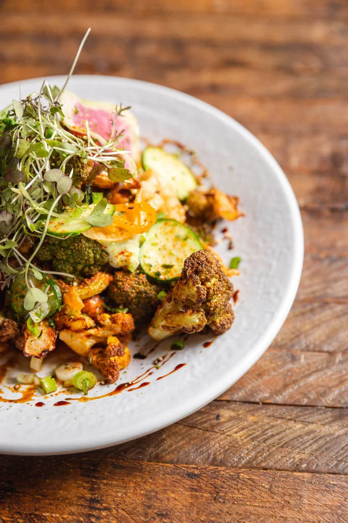 Plated roasted broccoli, cauliflower, zucchini, radish, and microgreens with sauce.