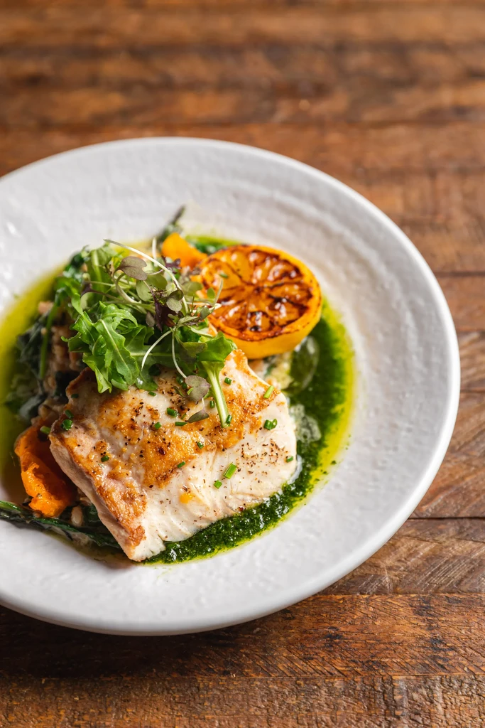 Plated seared fish, greens, charred lemon, and vibrant green sauce.