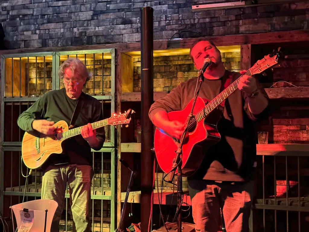 Two male musicians play guitars, one sings, under colorful lights.