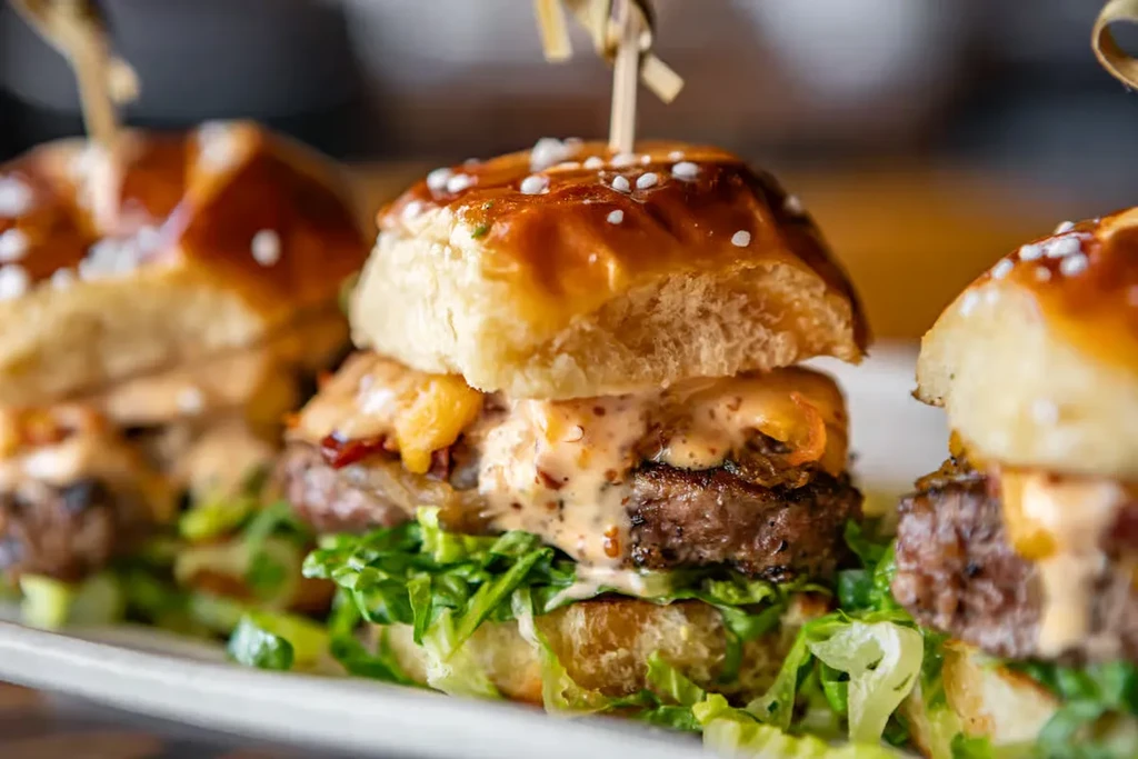 Three brioche sliders on a white plate, one skewered with sauce.