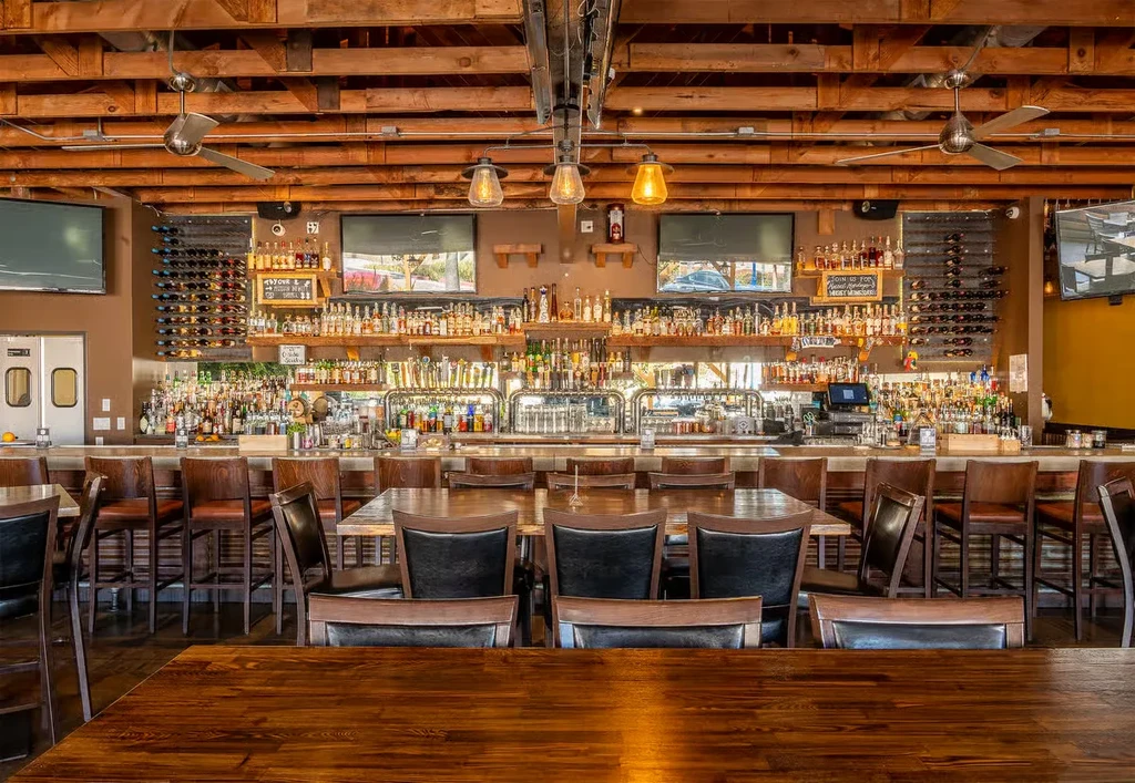 Modern rustic restaurant interior with a well-stocked bar and empty tables.
