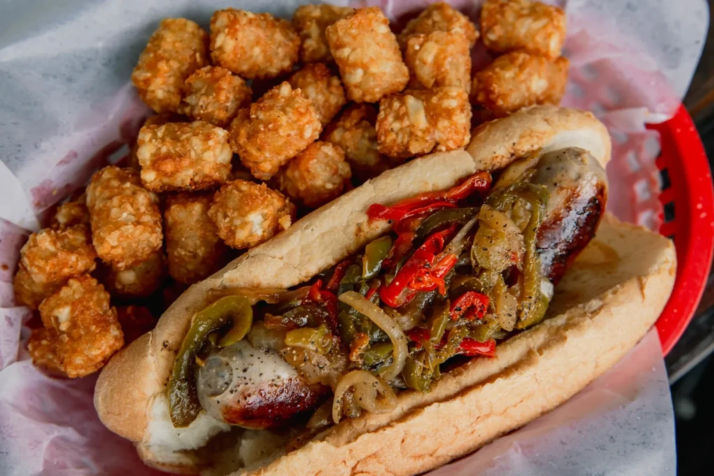 Grilled sausage with peppers, onions, and tater tots in a red basket.