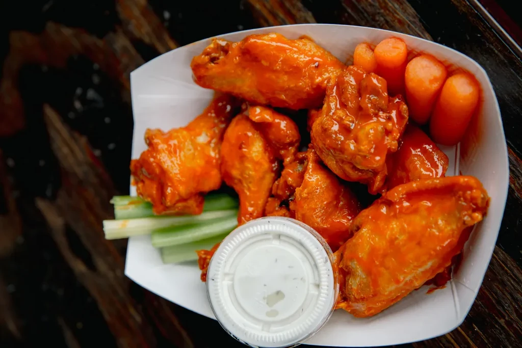 Buffalo wings, celery, carrots, and dipping sauce in a paper tray.