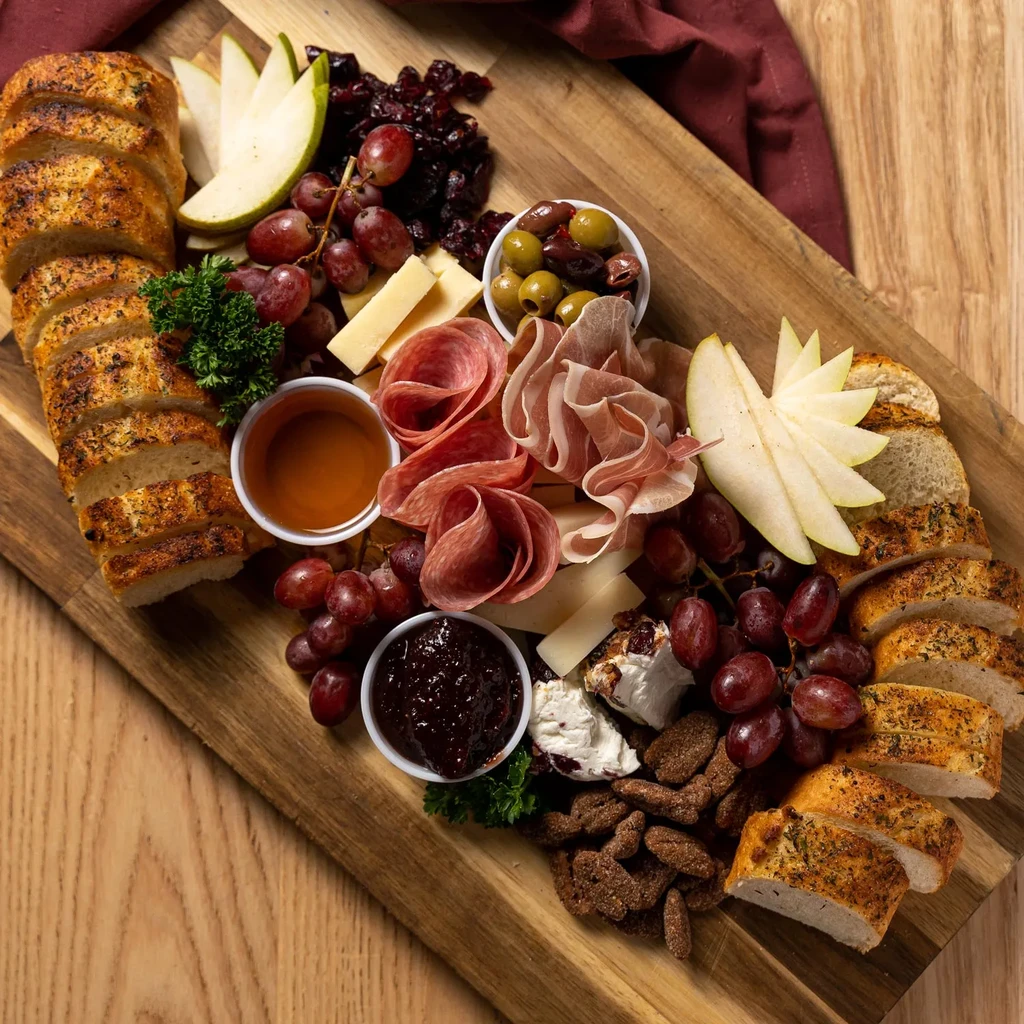 Large wooden charcuterie board displaying meats, cheeses, fruits, bread, and condiments.