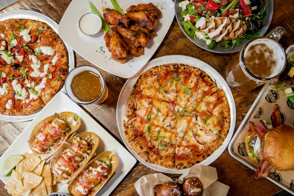 Diverse food spread: pizzas, tacos, wings, burger, salad, beer on a table.