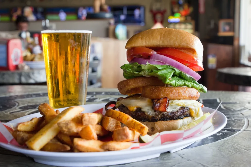 Stacked burger with fries and beer on a checkered paper plate.