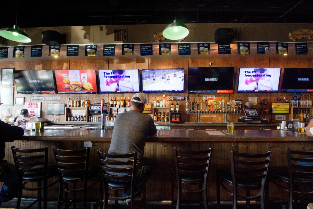 Bustling bar with a long wooden counter, many beer taps, and TVs.