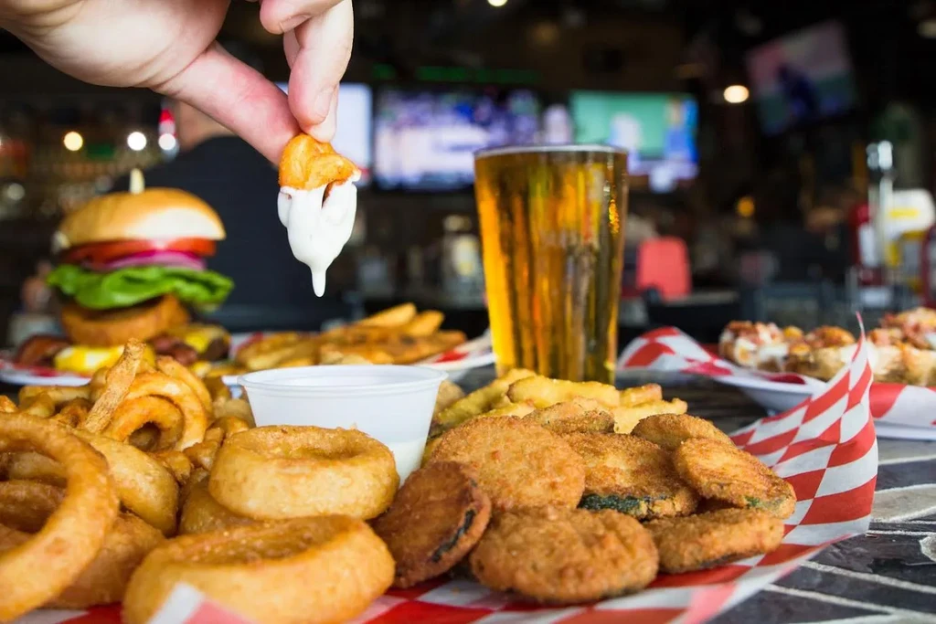Hand dips fried food from a platter, with beer and blurred sports TVs.