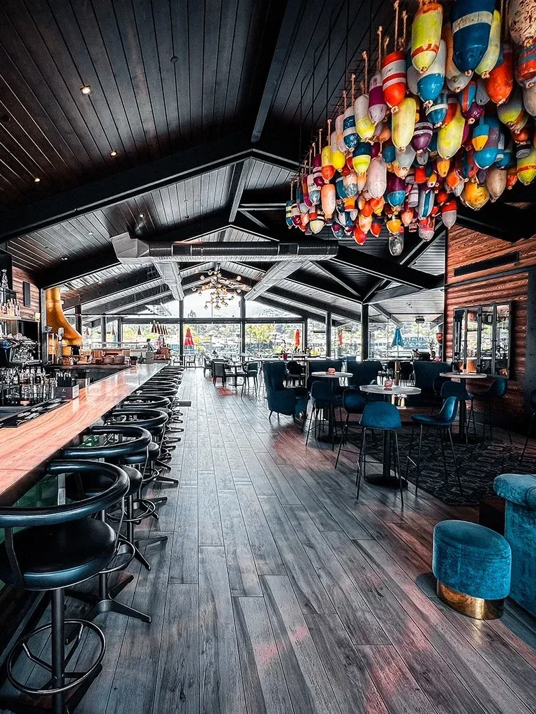 Restaurant interior with dark wood ceiling, light floor, and buoy chandelier.