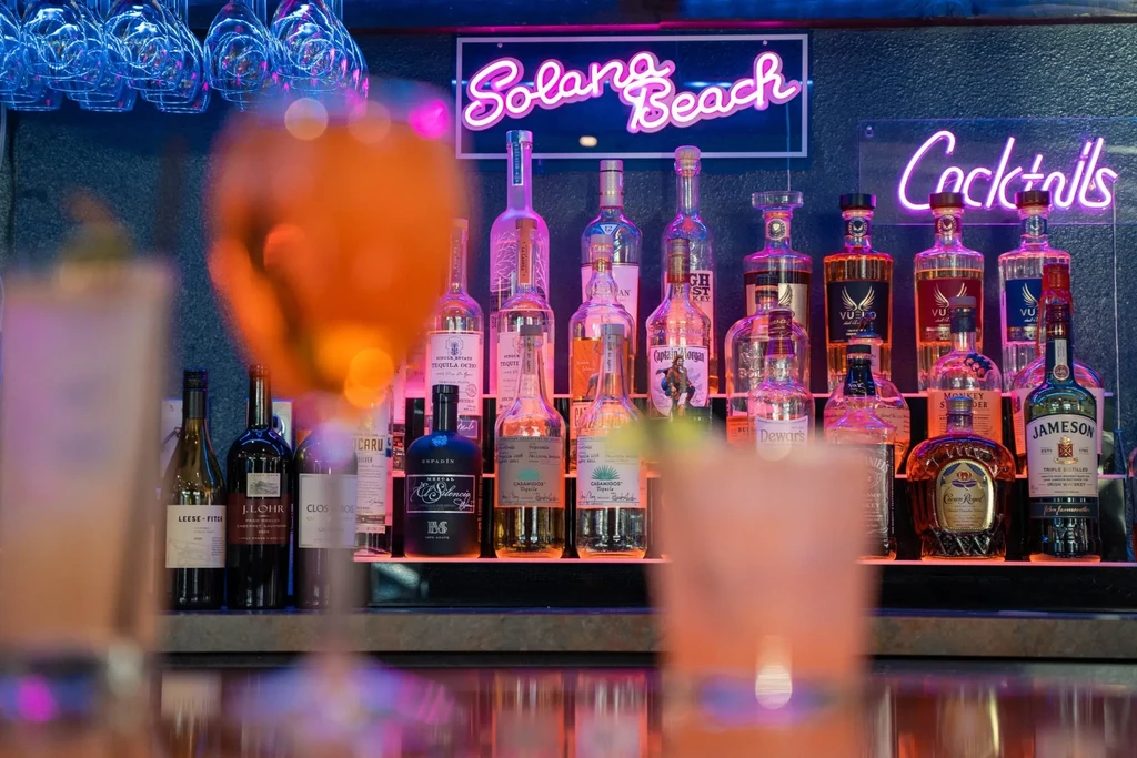 Brightly lit liquor shelves behind blurred cocktails, with pink neon signs.