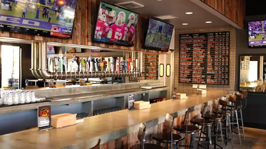 Modern sports bar with concrete counter, beer taps, and TVs showing games.