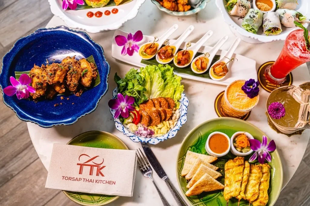 Overhead flat-lay of vibrant Thai dishes and cocktails, garnished with orchids.