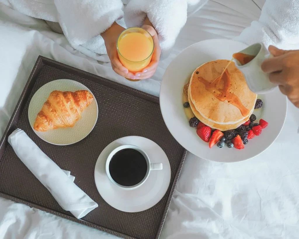 Overhead breakfast-in-bed: pancakes with berries, croissant, coffee, juice, pouring syrup.