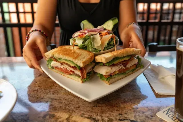 Hands present a hearty club sandwich and fresh green side salad.