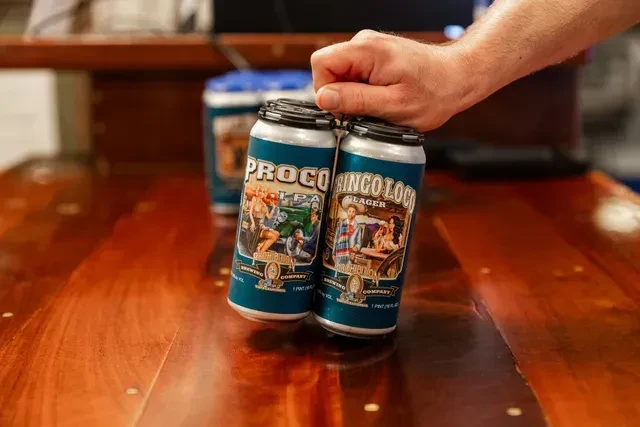 Hand holds two brightly illustrated beer cans, 'PROCO IPA' and 'RINGO LOCO LAGER'.