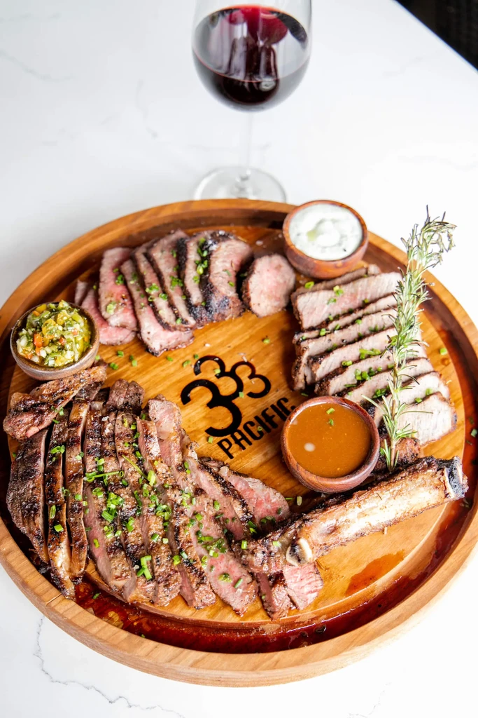 Medium-rare grilled steak, herbs, sauces, and wine on a branded board.