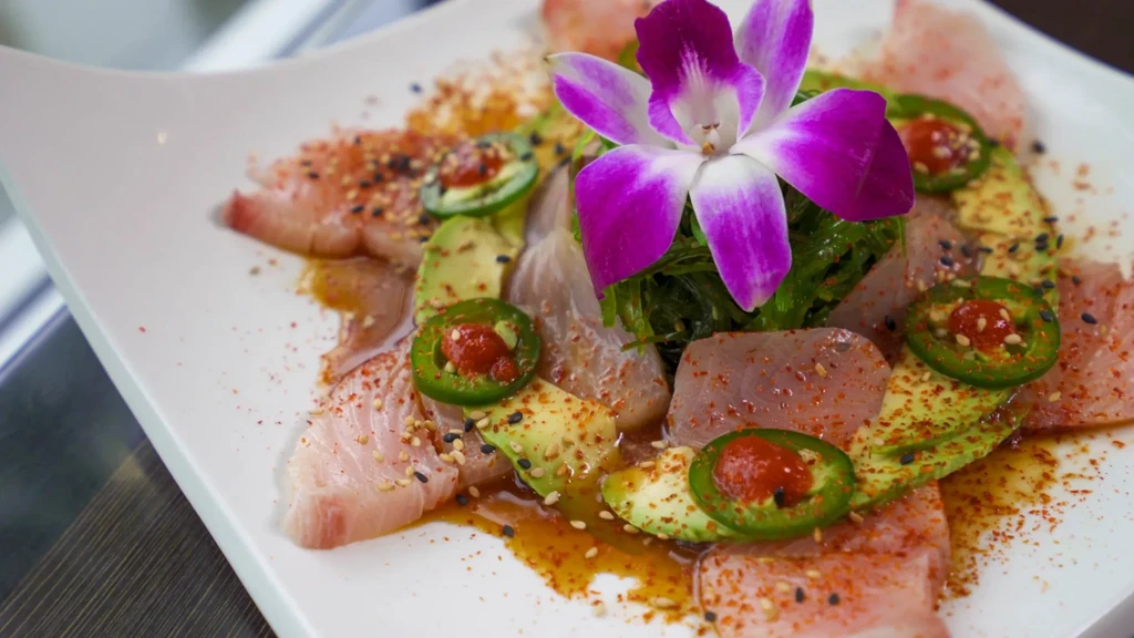 Thin-sliced fish dish with avocado, jalapeño, seaweed, and orchid garnish.