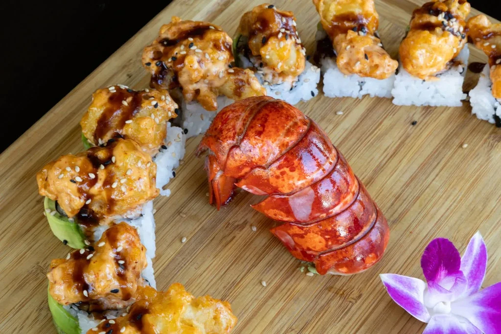 Sushi rolls with sauces, lobster tail, and orchid on wooden board.
